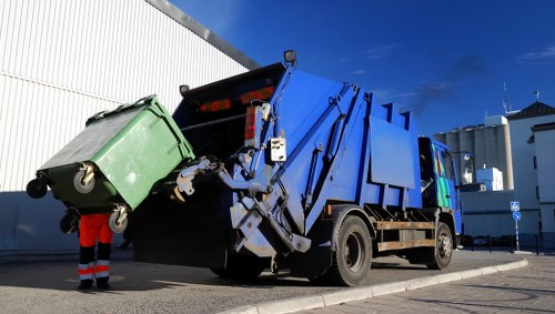 A large blue and black refuse collection truck is parked on a paved street next to a white industrial building with a corrugated exterior. The truck is actively emptying a green wheeled waste bin into its hopper; the bin is positioned near the rear of the vehicle and is supported on a metal frame with wheels visible at the base. The green bin appears to be made of hard plastic with a textured surface, and its lid is open, allowing the contents to be poured into the truck. The truck's hydraulic mechanism is partially visible, lifting and tilting the bin for waste transfer. A worker dressed in high-visibility red and black clothing is standing behind the bin, operationally assisting with the process. The sky above is bright blue with some wispy clouds, indicating clear weather. The surrounding environment suggests an urban or semi-industrial area, consistent with a rubbish removal service presence in the region near Hanwell, London, where Garden Clearance Hanwell operates. The scene highlights professional waste collection activity, aligned with rubbish disposal and clearance services essential for local maintenance.