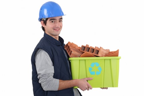 A young male waste disposal worker wearing a blue safety helmet and a navy-blue vest over a grey long-sleeved shirt, holding a bright green recycling bin with a blue recycling symbol on its side. The bin is filled with reddish-brown clay bricks and other construction debris, which are stacked neatly but protrude slightly at the top. The background is plain white, emphasizing the worker and the bin. This image visually represents rubbish collection and clearance services, suitable for a company like Garden Clearance Hanwell that specialises in garden and waste removal within the local postcode area, potentially serving properties in Hanwell and nearby districts. The scene highlights typical materials encountered in rubbish removal tasks, including small brick fragments, with a focus on eco-friendly waste segregation, depicted by the recycling bin. The professional attire and equipment suggest readiness for outdoor clearance work, aligning with service categories offered in the Hanwell area. The lighting is uniform, with no shadows, ensuring clear visibility of all objects and details, suitable for illustrating waste management services on a UK-based rubbish removal website.