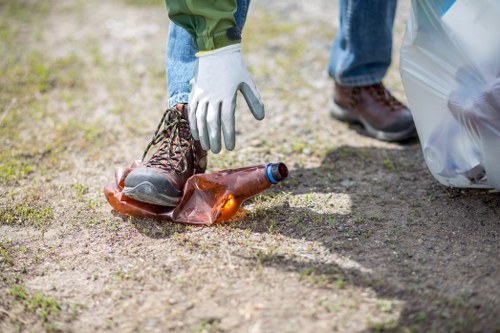 A close-up of a person's hand wearing a white work glove, reaching down to pick up a brown glass bottle from a patch of bare earth with a mix of soil and small grass patches. The individual is wearing blue jeans and brown work boots. In the background, part of another person's leg is visible, along with a large grey plastic rubbish bag. The scene appears to be outdoors, possibly in a garden or on a driveway, reflecting a rubbish removal or clearance activity. The image emphasizes cleaning up and removing waste material, which aligns with services provided by Garden Clearance Hanwell for rubbish collection and clearance tasks.