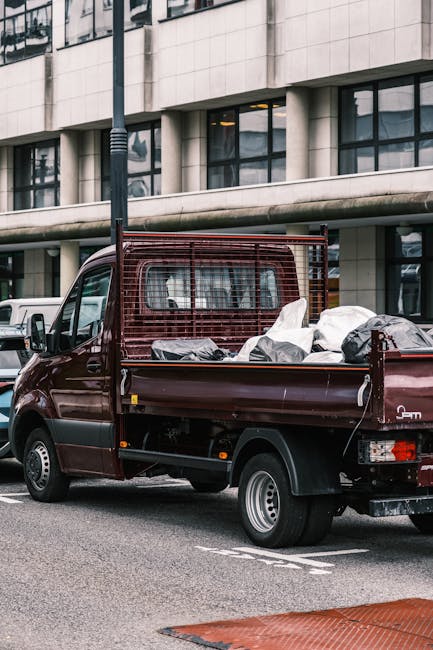 A dark red flatbed truck parked on a city street in front of a modern office building with large, reflective glass windows and a concrete facade. The truck's cargo area is filled with various waste ma