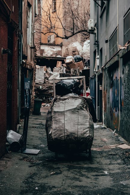 A pile of mixed waste materials, including cardboard boxes, paper, and plastic packaging, is realistically depicted stacked against a rough stone wall with a brick section in the background. The cardb