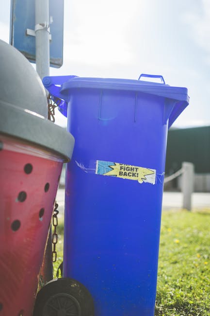 A large green plastic waste bin filled with several unopened bags of garden soil or compost, positioned on a paved surface against a dark blue brick wall. The bags are red with white text detailing pr